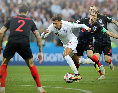 Футбольный матч между сборными Англии и Хорватии. Игрок в белой форме пытается продвинуться вперёд, обходя соперников в чёрно-красной форме. На заднем плане трибуны с болельщиками