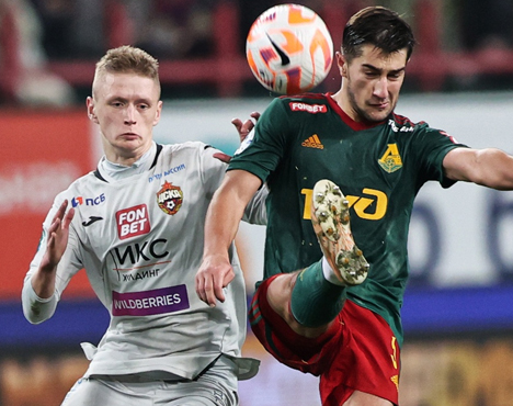 Два футболиста в моменте игры: один в серой форме тянется к мячу, другой в тёмно-зелёной форме соревнуется за мяч высоко поднятой ногой. Они играют на футбольном поле во время матча