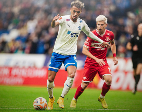 Футболист в бело-синей форме ведет мяч на поле, преследуемый игроком в красной форме. На заднем плане видно размытое изображение зрителей на трибуне