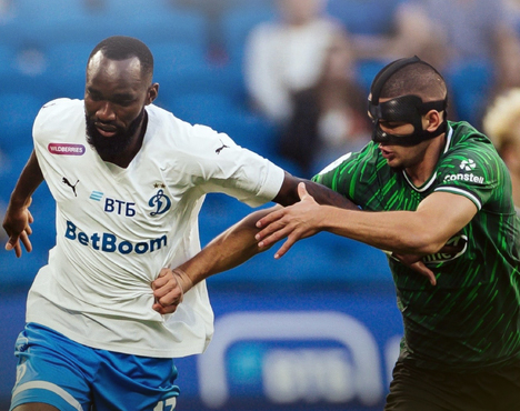 Футболист в белой форме команды Динамо с мячом на поле, в то время как соперник в зелёной форме пытается его остановить. Соперник носит чёрную маску. На заднем плане видна размытка трибун