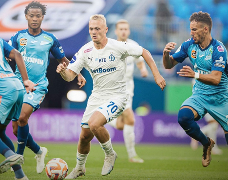 Футбольный матч: игрок в белой форме с мячом движется в окружении игроков в голубой форме. Яркие цвета формы, динамичное действие на зелёном поле