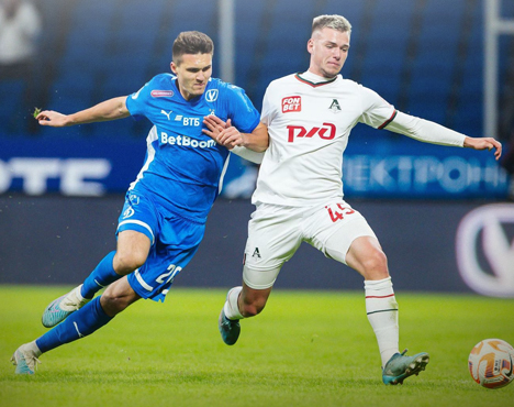 На изображении двое футболистов соревнуются за мяч на поле. Игрок слева в синей форме, а справа - в белой. Оба активно борются, выражая решимость и концентрацию. Виден стадион с надписями на фоне