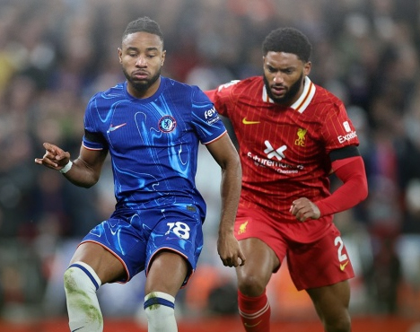 Двое футболистов на поле в динамичном моменте игры. Один одет в синий комплект формы, другой в красный. На заднем плане видны размазанные силуэты зрителей