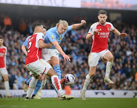 На изображении футболисты в разгаре матча: игрок в голубой форме пробивает мяч, противодействуя двум игрокам в красно-белой форме. На заднем плане видны зрители на трибунах