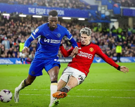 На изображении два футболиста на поле во время матча: один в синей форме, другой в красной. Игрок в синей форме устремлён с мячом вперёд, а игрок в красной пытается помешать, выполняя подкат. Стадион заполнен зрителями