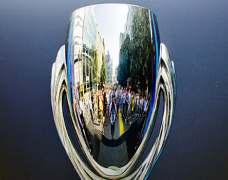 На изображении отражение городской сцены с людьми и зданиями в поверхности полированного серебристого предмета, напоминающего трофей или кубок, на нейтральном темном фоне