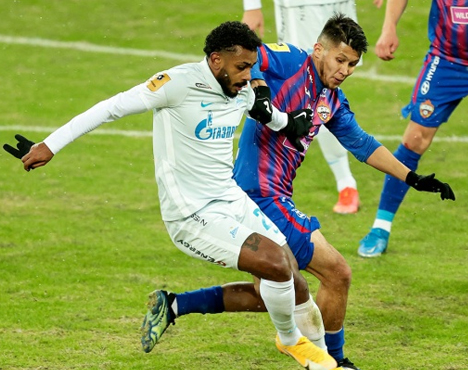 На изображении два футболиста в динамичной игре. Один в белой форме, другой в сине-красной. Они соревнуются на поле, борясь за мяч. Видны детали стадиона и окружающая обстановка спортивного матча