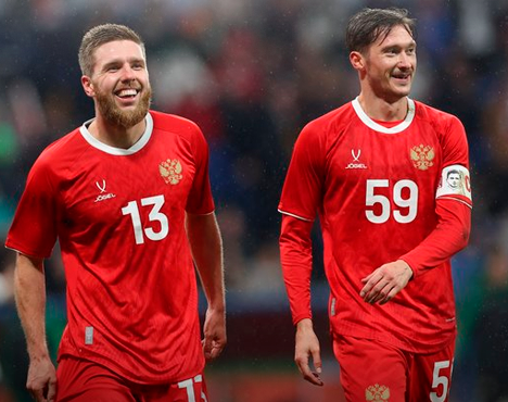 Двое футболистов в красной форме сборной России с номерами 13 и 59 на футболках, стоят на поле. На одном из игроков повязка капитана. Фон размыт, предположительно стадион