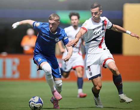 Два футболиста в моменте игры: игрок команды в синей форме ведёт мяч под прессингом соперника в белой форме. На дальнем плане видны размытие трибун и игроков