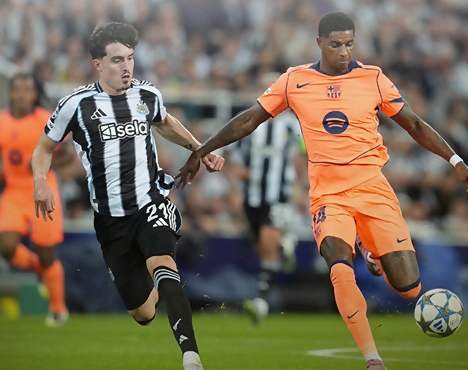 На изображении два футболиста борются за мяч на поле во время матча. Один из них одет в черно-белую форму, другой — в ярко-оранжевую. На заднем плане видны зрители и другие игроки