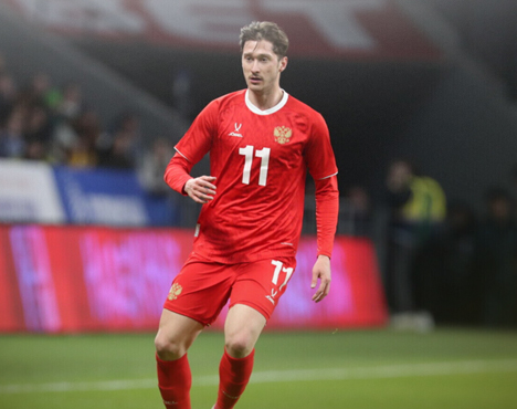 Футболист в красной форме с номером 11 играет на поле. На заднем плане размытый стадион и болельщики